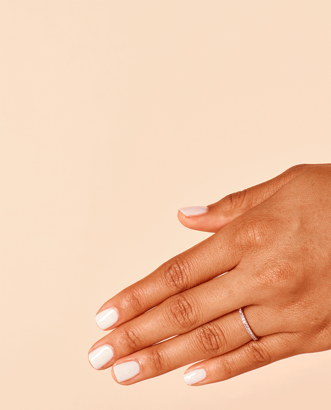 Hand with short, glossy white nails painted in Funny Bunny® nail lacquer rests against a peach background, showcasing the soft neutral shade as part of an OPI nail polish gift set idea for everyday wear.