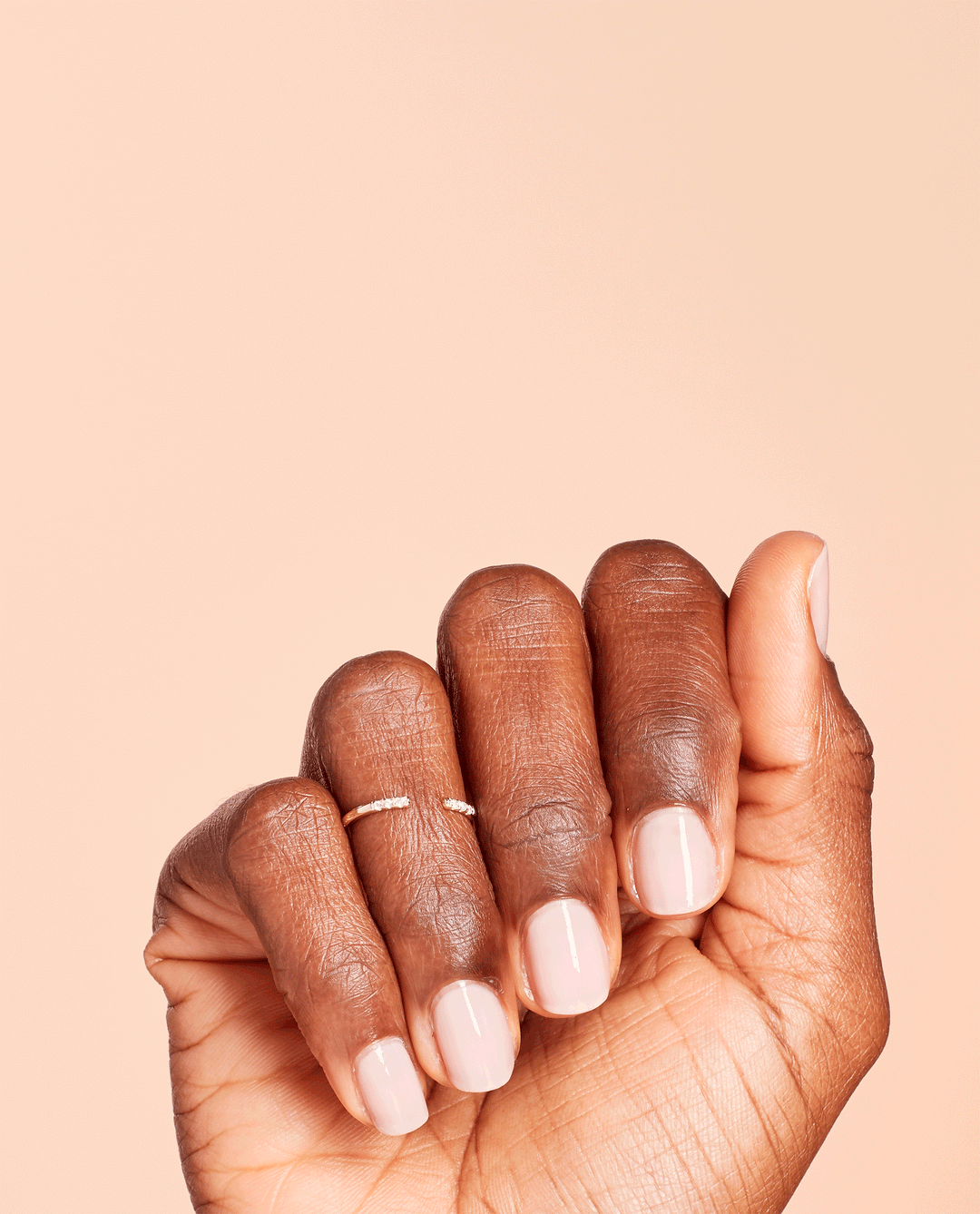 Hand with short, neatly manicured nails wears OPI Put it in Neutral nail lacquer, showing a soft pink nude shine against a peach background, ideal for promoting an elegant everyday OPI nail polish gift set in the US.